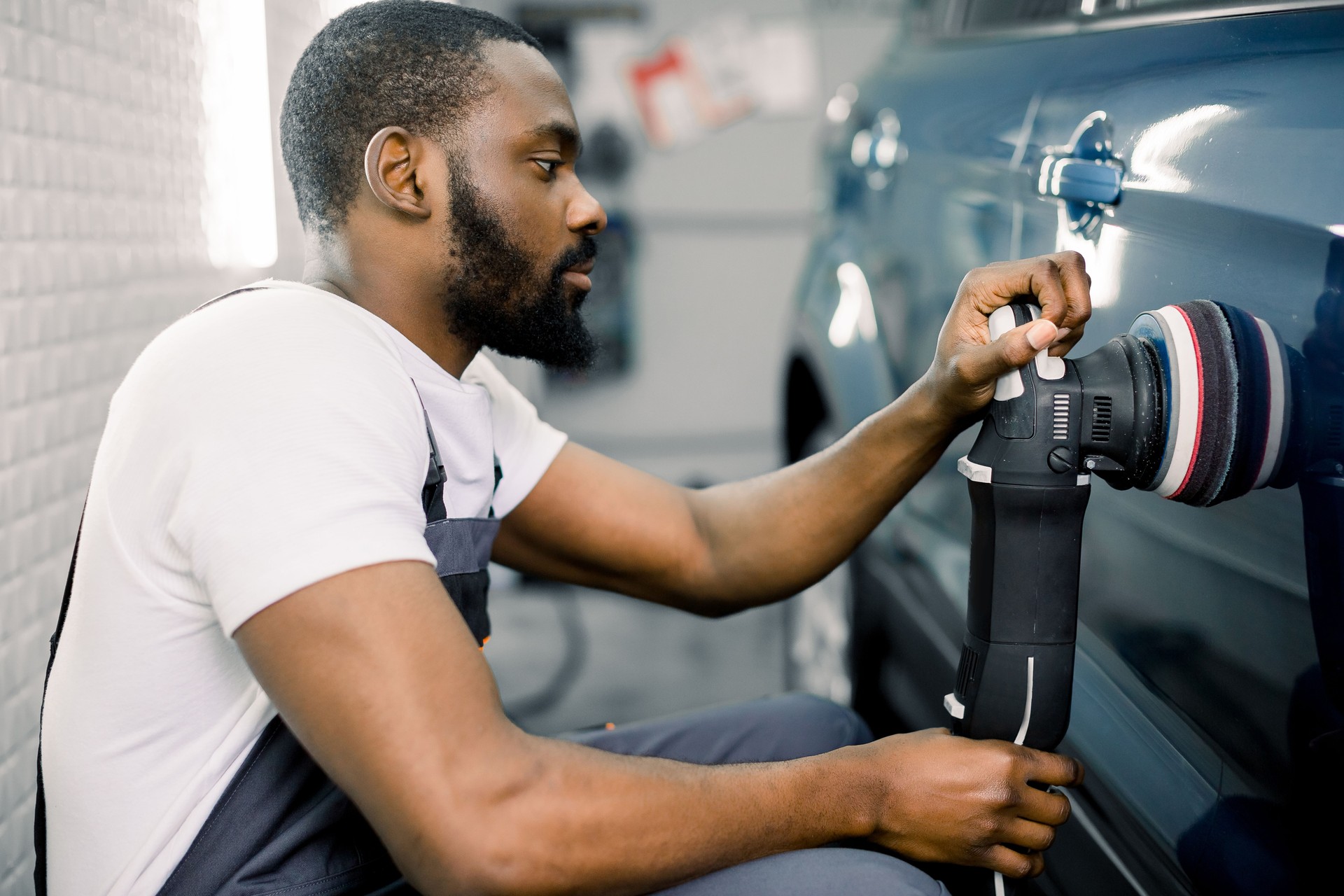 Auto detailing service, polishing of the car. Side view of young African American man worker n t-shirt and overalls, polishing blue car door with orbital polisher