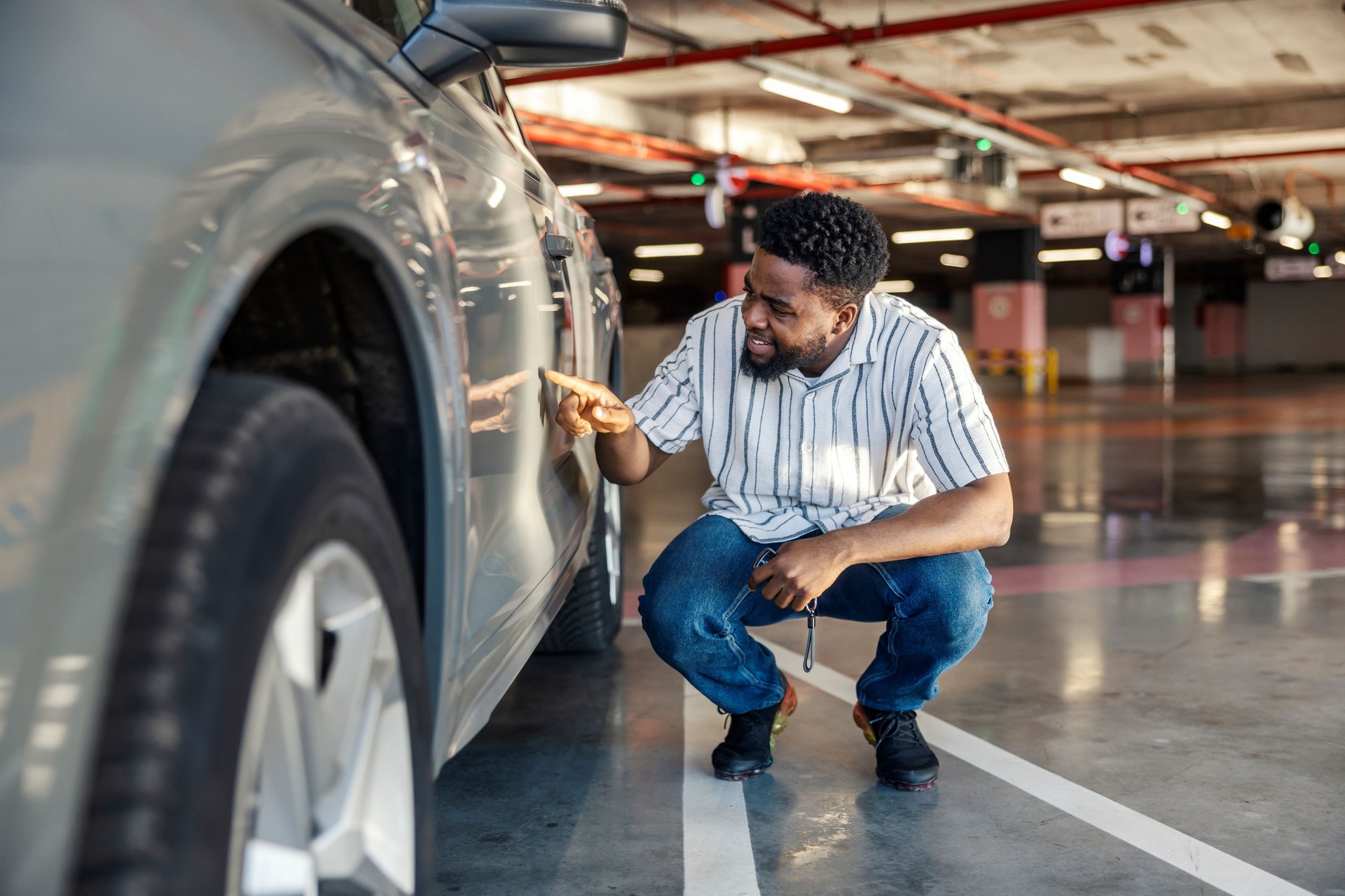Black guy is crouching next to his car and looking at little scratch on car door.